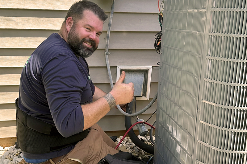Technician giving thumbs up while working on an outdoor HVAC unit next to a house with beige siding.