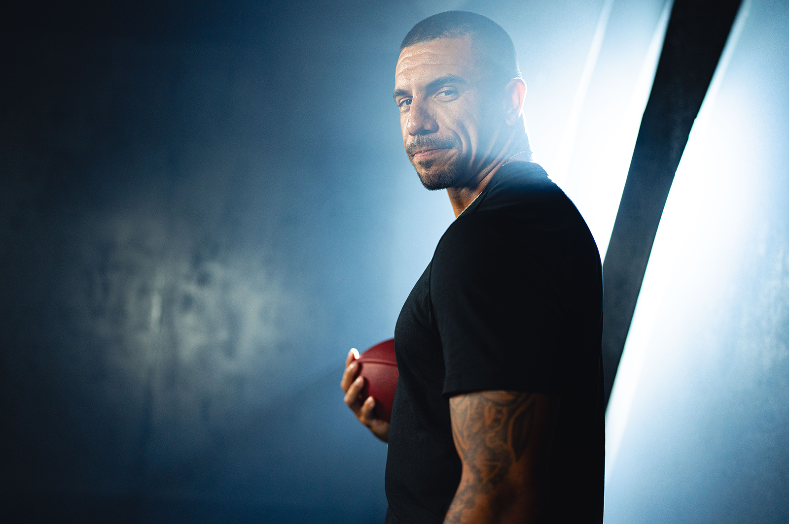 Man with tattoos holding a football, looking over his shoulder against a dramatic blue-lit background.