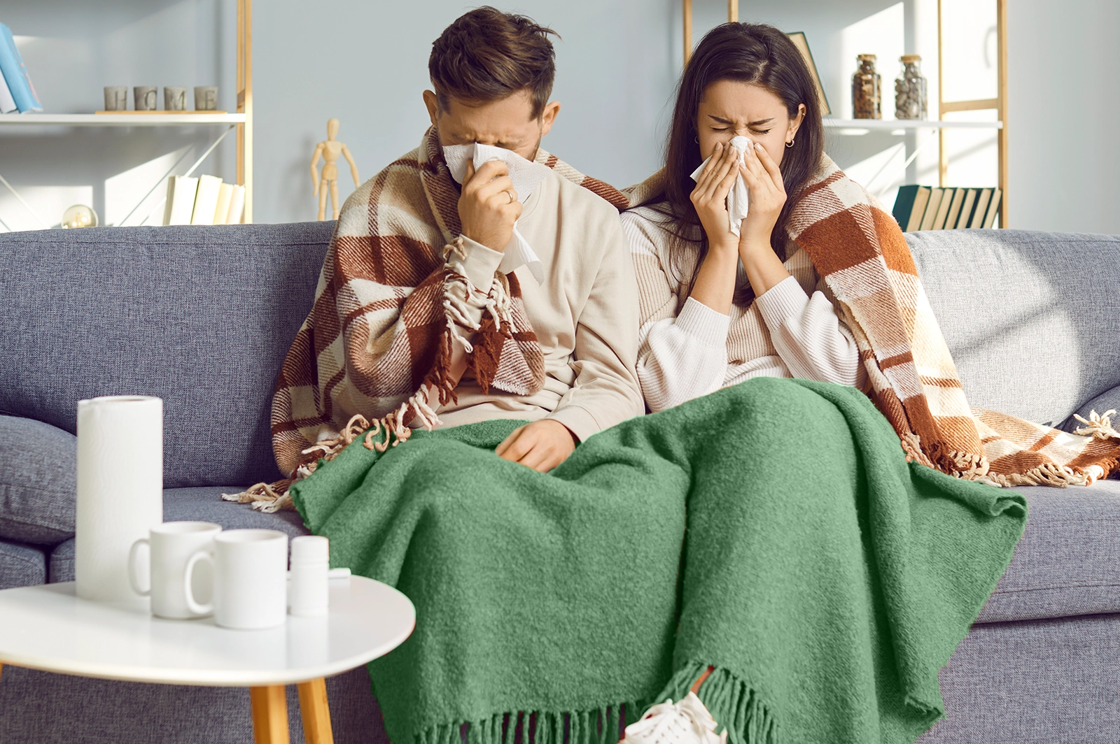 Two people sit on a couch under a blanket, both using tissues to blow their noses. A table in front holds cups, medicine bottles, and a roll of paper towels.