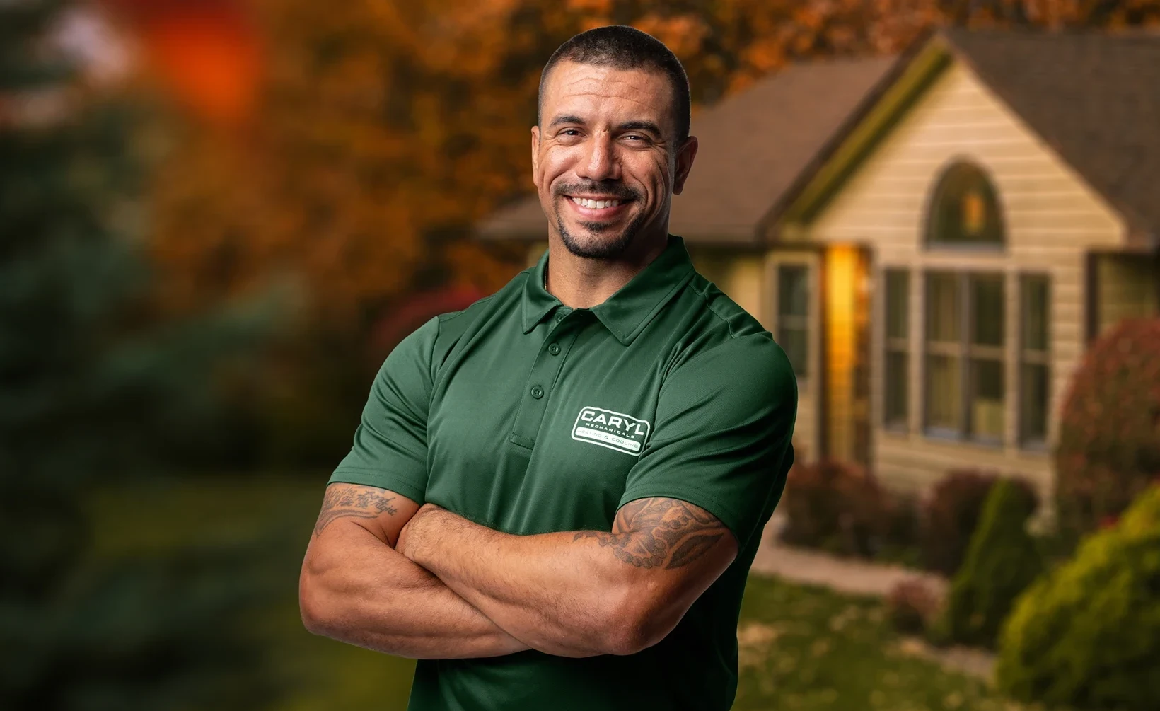 Man in a green polo shirt with crossed arms stands smiling in front of a house with autumn trees in the background.