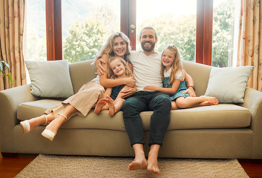 A family of four sits closely together on a beige sofa, smiling and posing for the camera in a bright living room with large windows.
