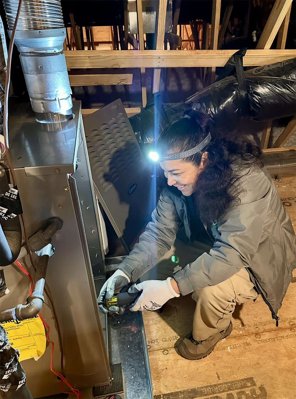 Person wearing gloves and a headlamp repairs a furnace in an attic. Tools and wiring are visible, and the furnace panel is open. Wooden beams and insulation appear in the background.