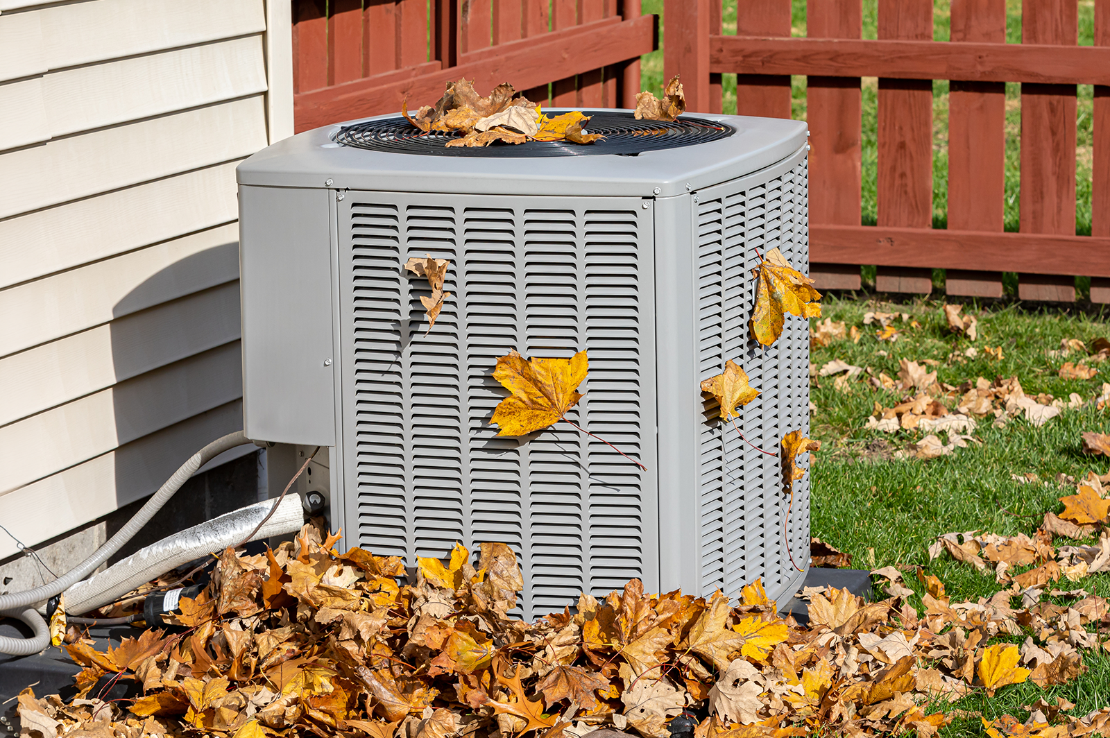 A grey outdoor air conditioning unit covered in fallen autumn leaves.